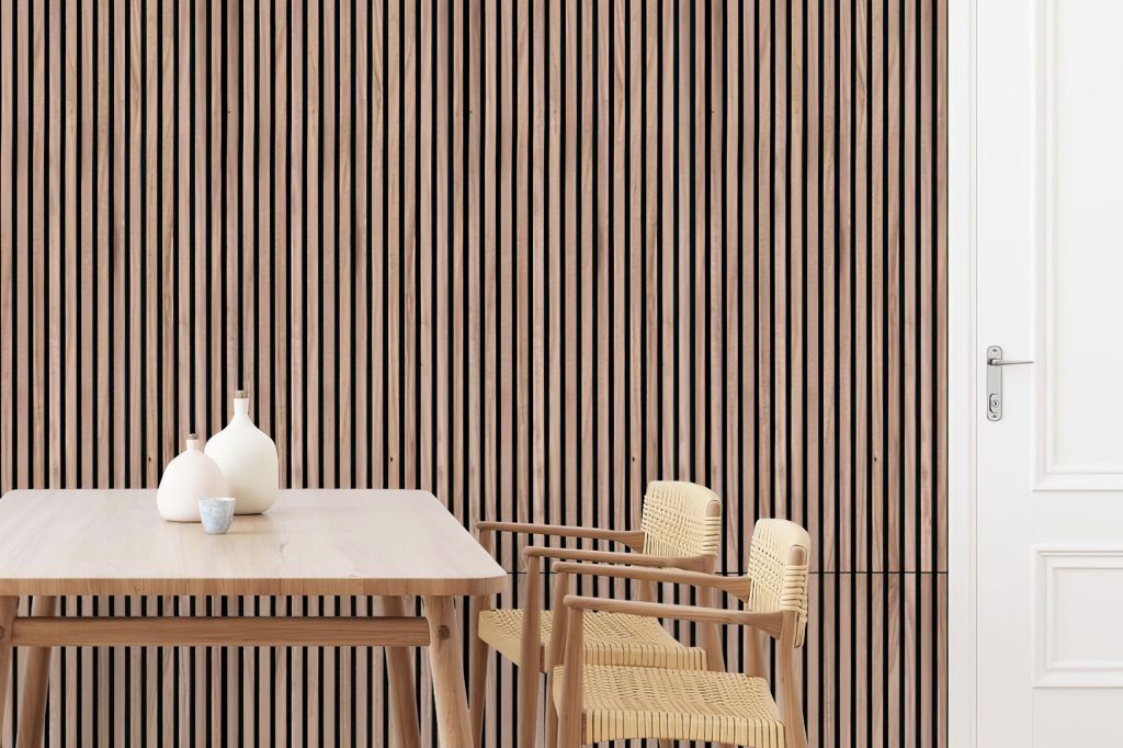 Minimalist dining area with a light wood table, two woven chairs, and decorative white vases. The background features Stilj™ Acoustic Wood Panel Installations as a modern vertical slat wall next to a white door with a silver handle.