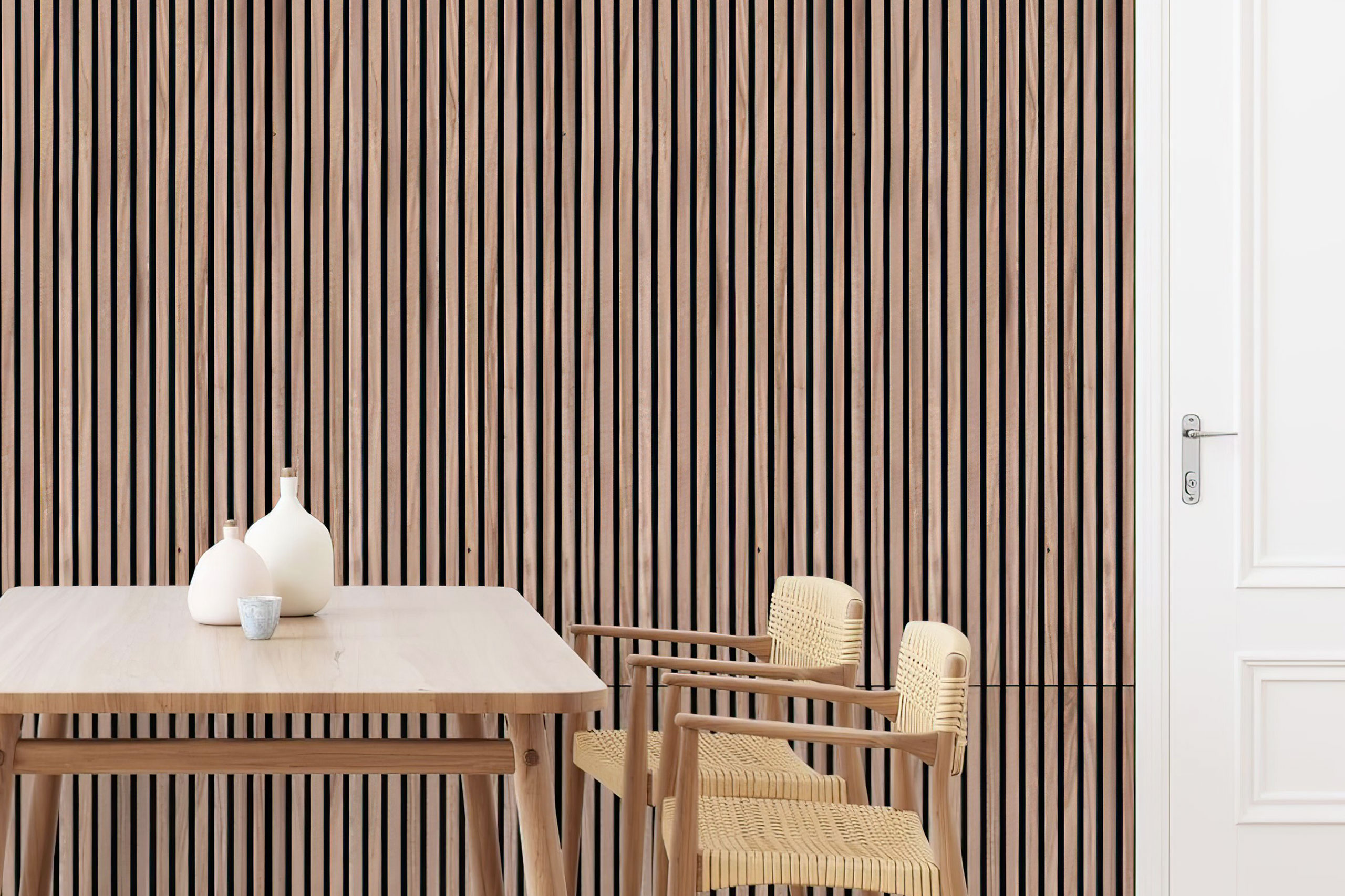 Minimalist dining area with a light wood table, two woven chairs, and decorative white vases. The background features Stilj™ Acoustic Wood Panel Installations as a modern vertical slat wall next to a white door with a silver handle.
