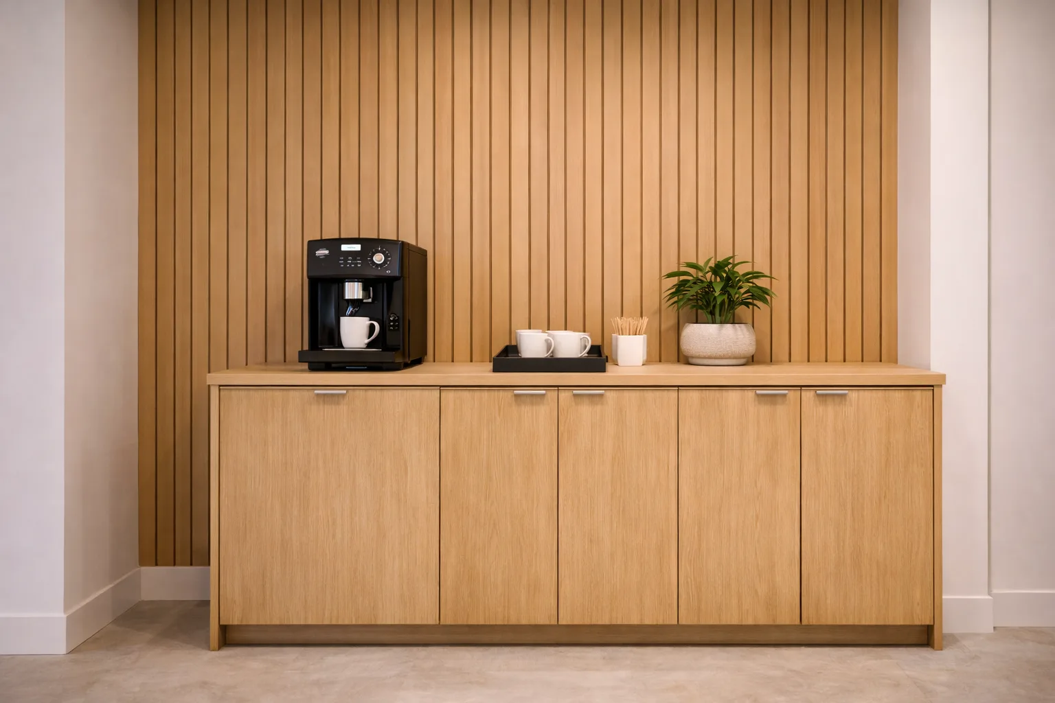 A modern coffee station with a black coffee machine, two white cups, a tray with stirrers, and a potted plant on a light wood cabinet against a wooden slat wall.