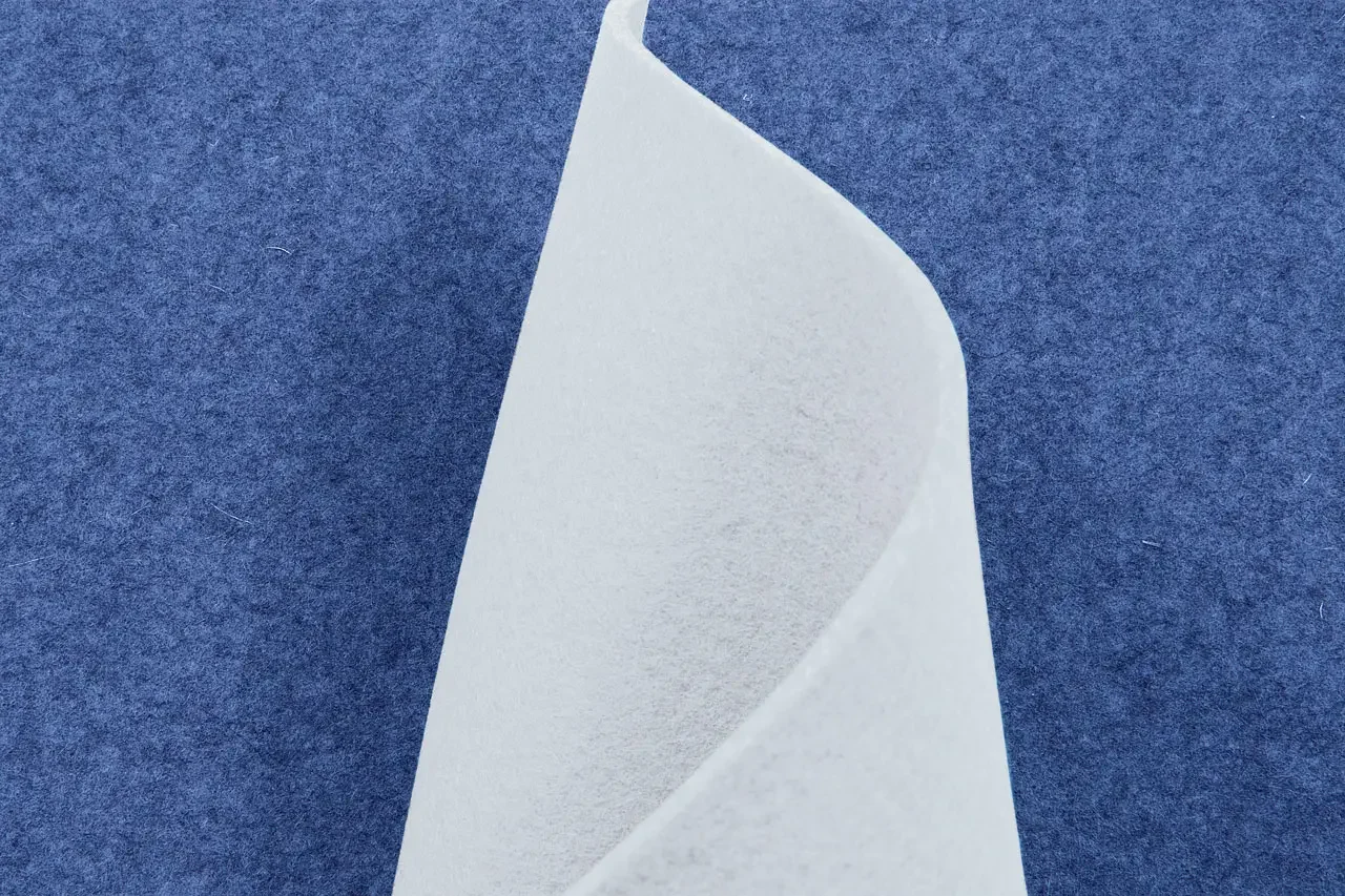 A close-up of a single white sheet of paper with a curled corner, set against a textured blue background featuring Stilj™ Sustainable Acoustic Wood Paneling.