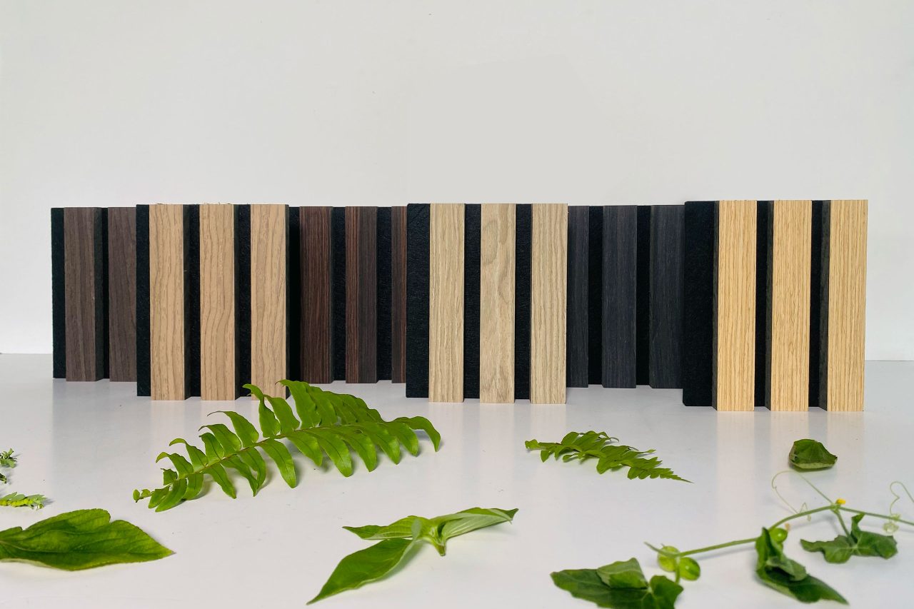 Rectangular wood samples in various shades of brown and black, resembling timber slat acoustic panels, are arranged upright in a row on a white surface with green leaves and small plants scattered in front. The background is plain white.