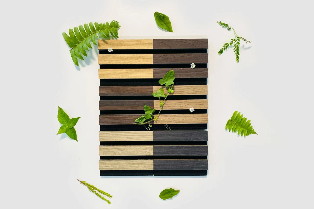 A square panel of Stilj™ Sustainable Acoustic Wood Paneling features alternating light and dark slats, adorned with small green plants and white flowers, all surrounded by various green leaves on a light background.