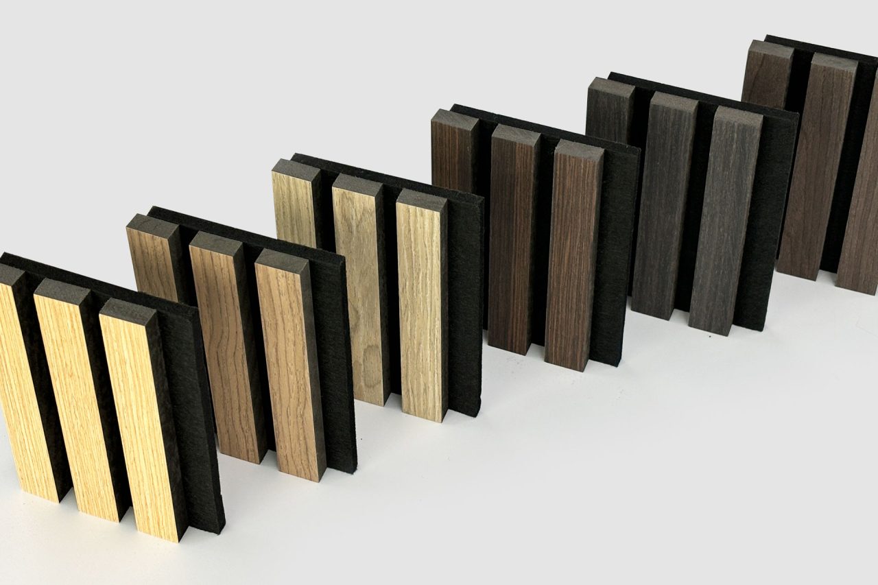 Several pairs of rectangular Stilj™ Acoustic Timber Slats in varying shades from light to dark are standing upright in a staggered row on a white surface.